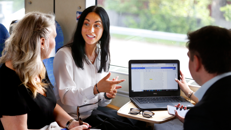 Tre personer som sitter på ett tåg och pratar, det finns en dator i mitten av dem.