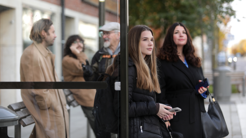 Fem personer står och väntar på bussen vid en busshållsplats. 