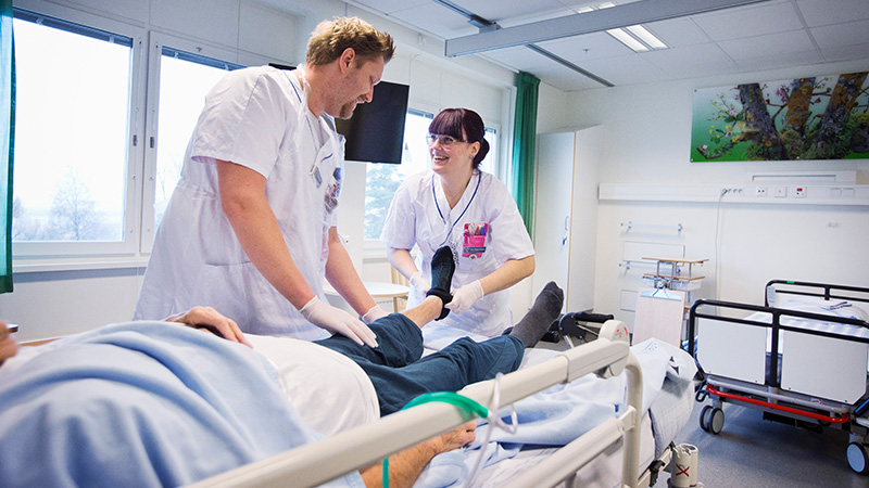 En manlig och en kvinnlig undersköterskor tittar på varandra under tiden de hjälper patient.