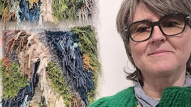 Ett färgglatt textilkonstverk med långa trådar hänger till vänster i bild. Till höger ser vi en leende kvinna med svarta glasögon som är nära i bild.