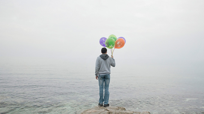 En person står vid en strand med ryggen vänd mot betraktaren och blickar ut över havet. Hen håller färgglada ballonger i ena handen. Omgivningen är disig och grå.