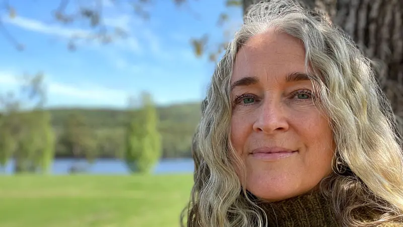 Kvinnoansikte med skog, äng och vatten i bakgrunden.