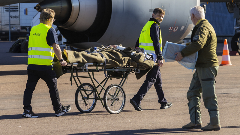 Två personer i gula varselvästar rullar en bår. På båren ligger en patient. I förgrunden syns en person som vänder sig om och tittar mot båren.