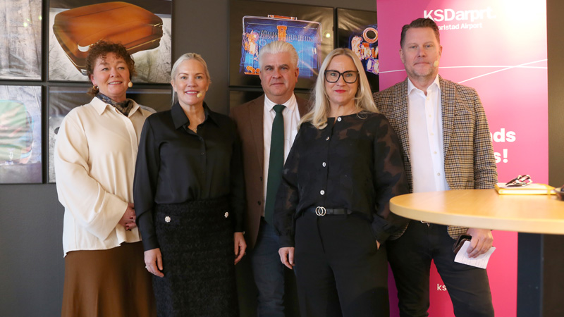 Fem personer står uppradade efter en pressträff. I bakgrunden syns en rosa roll-up och på väggarna hänger färgglada bilder.