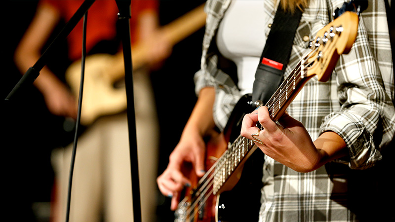 En person spelar gitarr och en annan person som spelar bas i bakgrunden. 