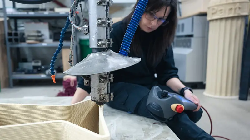 A woman sits on the edge of a bench, monitoring a machine that cuts out shapes.