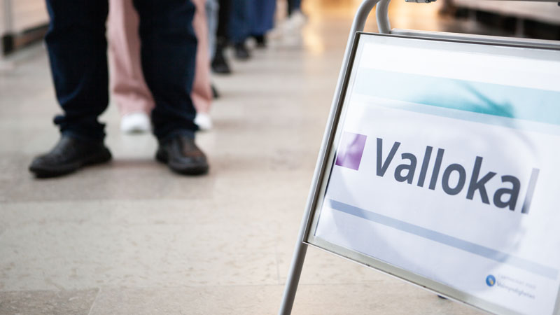En skylt med texten vallokal står på golvet. I bakgrunden syns fötterna på flera personer som står i kö.
