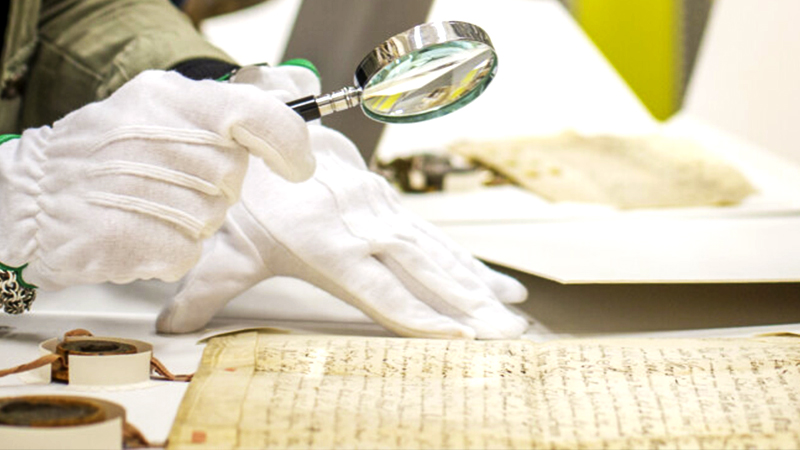 Händer med handskar på hanterar förstorningsglas och ett gammalt dokument.
