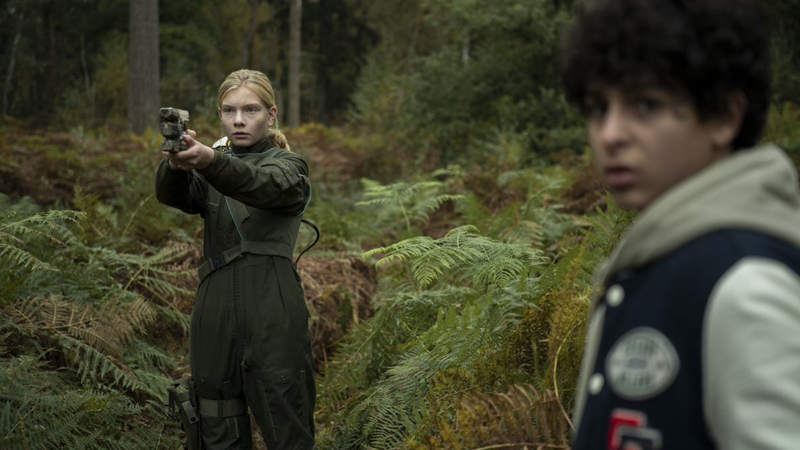 En flicka och en pojke i en skog. Flickan håller i ett vapen.