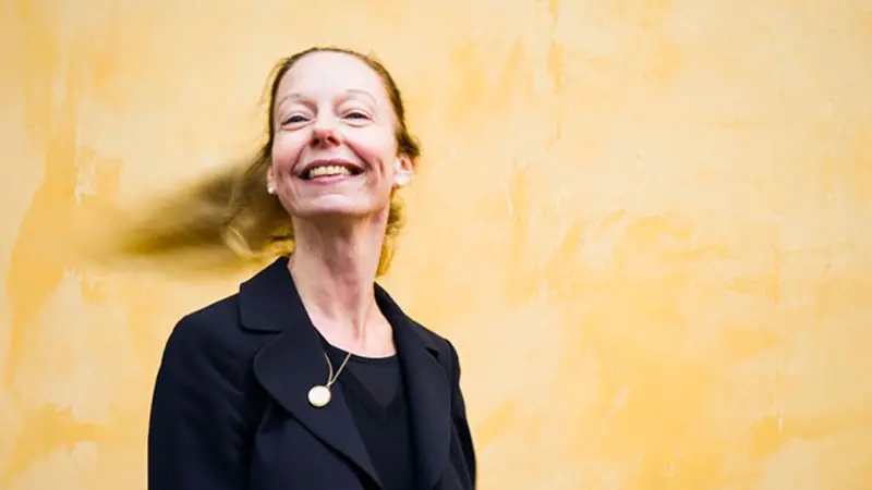 Kvinna i mörk kavaj står framför gul vägg, med hår som blåser åt sidan.