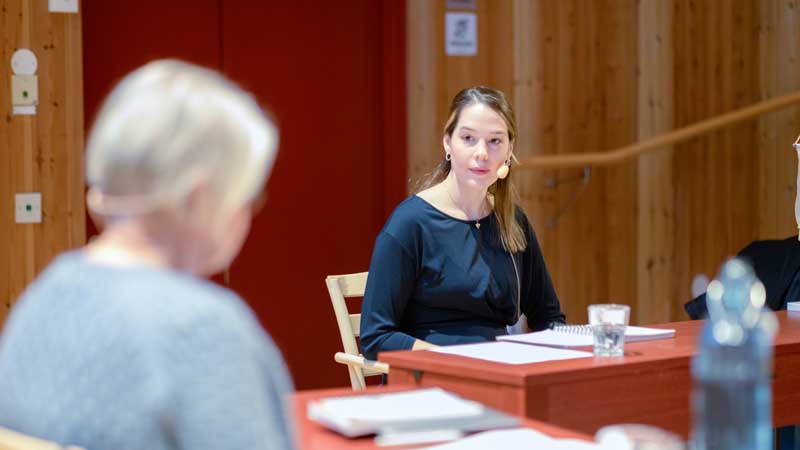 Noelle Probert sitter vid ett bord i stora salen på Värmlands museum. I förgrunden syns ryggen på en annan person.