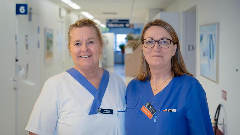 Två medarbetare i hälso- och sjukvårdens blåvita kläder. Bilden är tagen i en sjukhuskorridor.