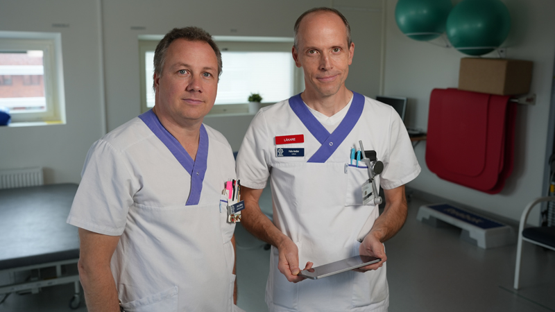 Två medarbetare i hälso- och sjukvårdens blåvita kläder. En av dem håller i en ipad.