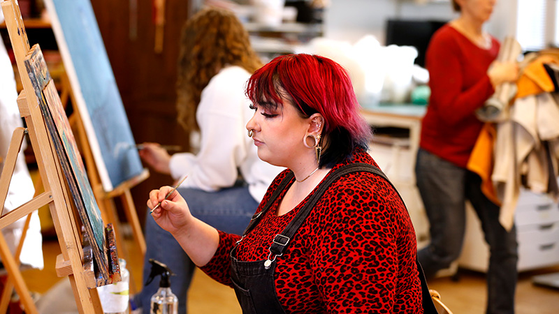 Kvinna i rött hår och röd tröja sitter framför ett staffli och målar, I bakgrunden syns andra studenter.