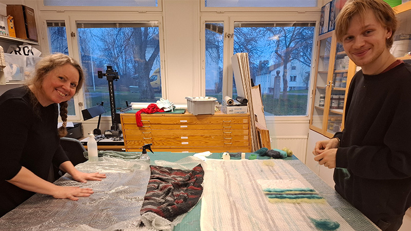 Kvinna och man ler mot kameran framför en stor bänk med textilier de arbetar med.