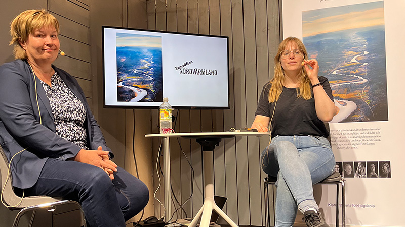 Två personer sitter på en liten scen med en skärm bakom sig på bokmässan. 