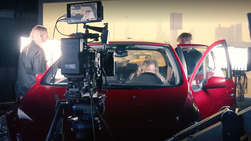 En kamera står fram för en röd bil i en studio med en stadsmiljö och trafik som bakgrund. 