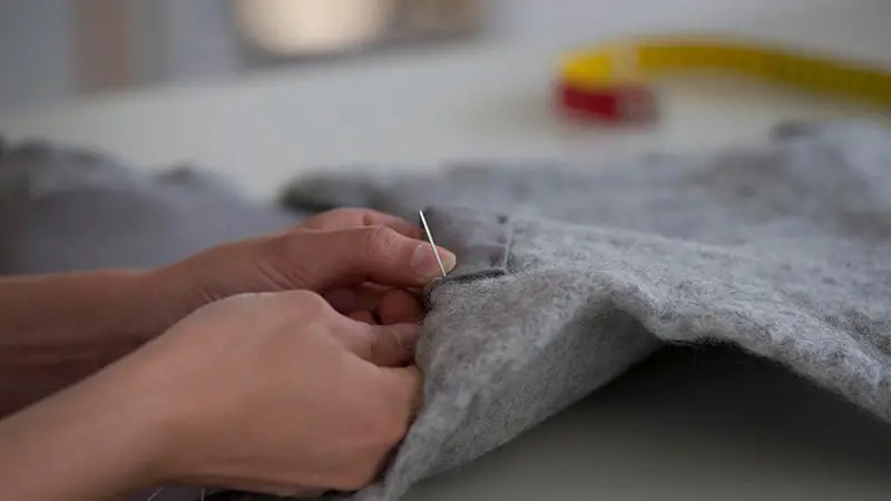 Närbild på händer som syr i grått ylletyg.