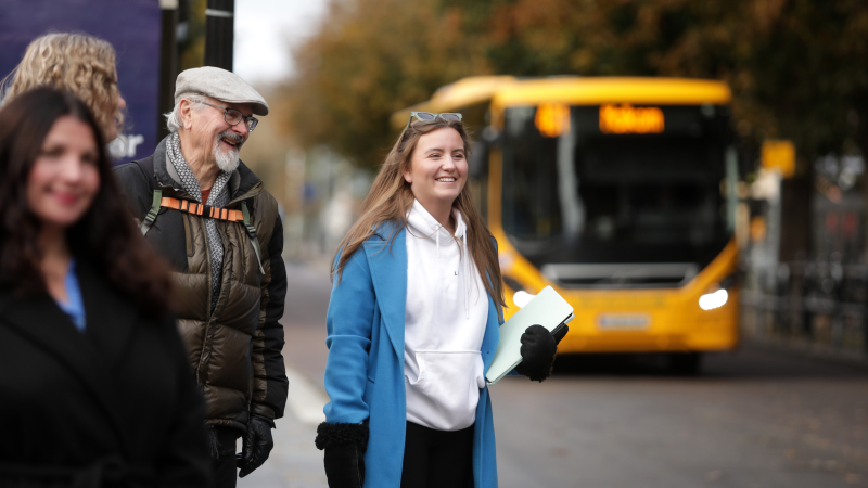 Personer väntar vid en busshållplats medan en gul buss närmar sig i bakgrunden.
