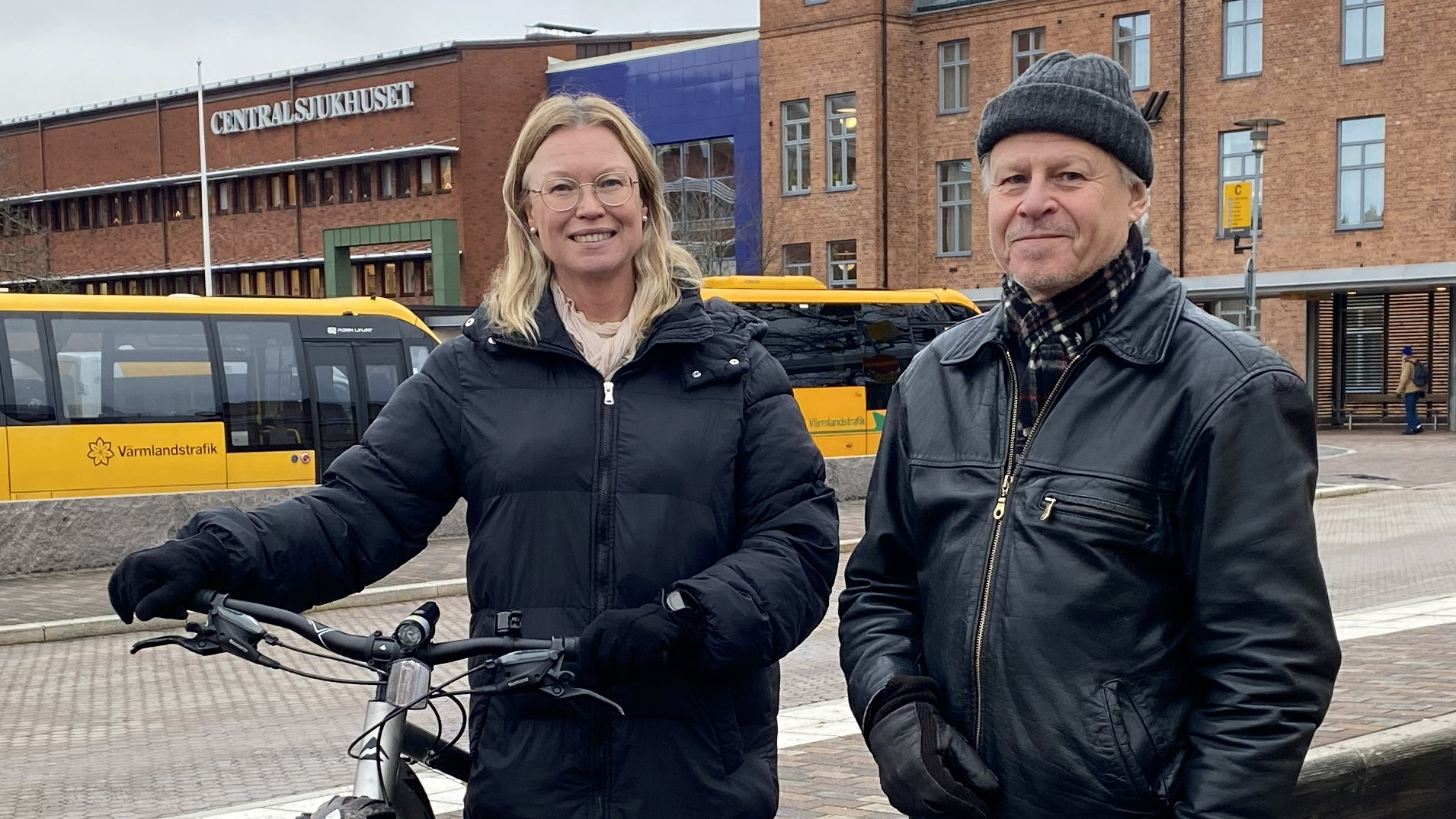 En kvinna och en man står utomhus och tittar in i kameran. Kvinnan håller i en cykel. Bakom dem syns gula bussar.och en byggnad med texten Centralsjukhuset.