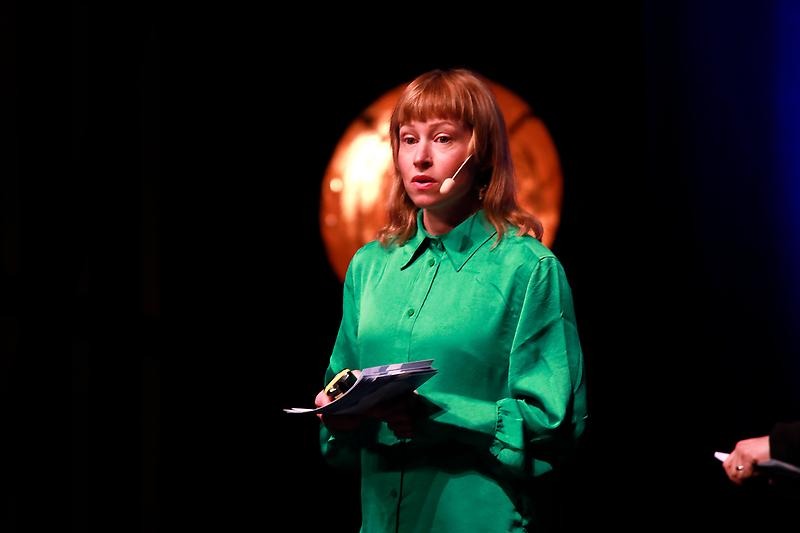 Linda Moestam. En person i grön skjorta står på en scen.