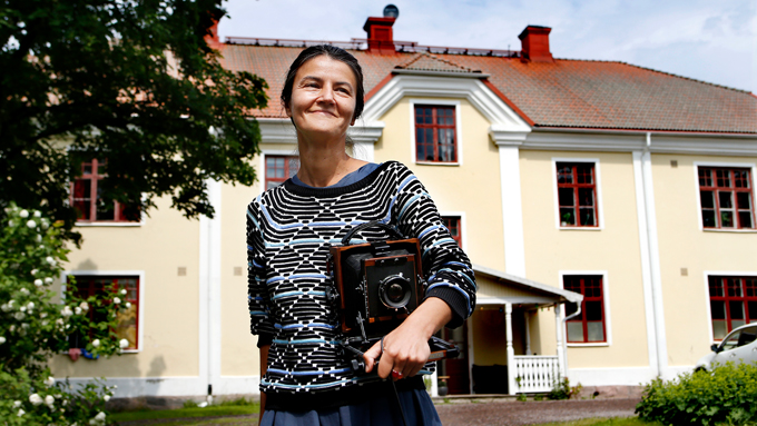 Foto av konstnären Mila Teshaieva  framför Gunnerudsgården. Mila ler och håller i en analog kamera.