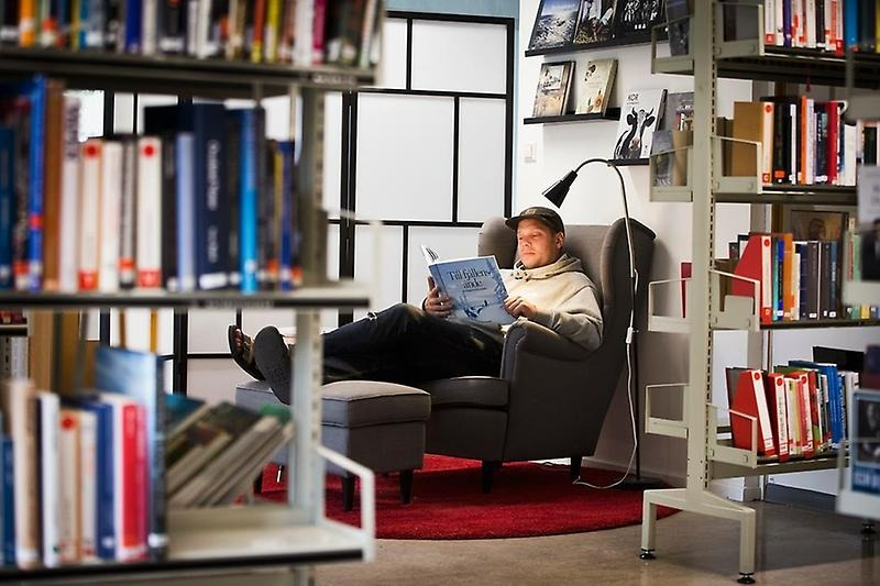 Person sitter i en fåtölj i ett bibliotek och läser en bok