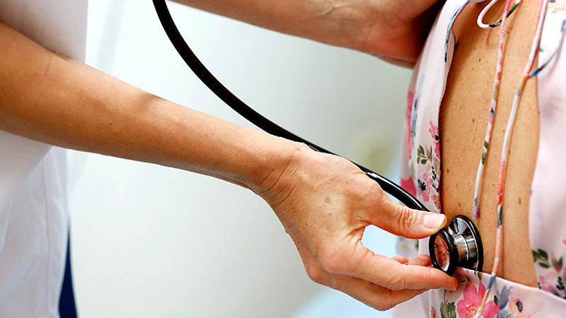 En läkare lyssnar på en patient andningsvägar genom ett stetoskop tryckt mot ryggen.