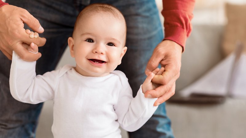 Litet barn ler i kameran. En vuxen persons händer håller i barnets händer.
