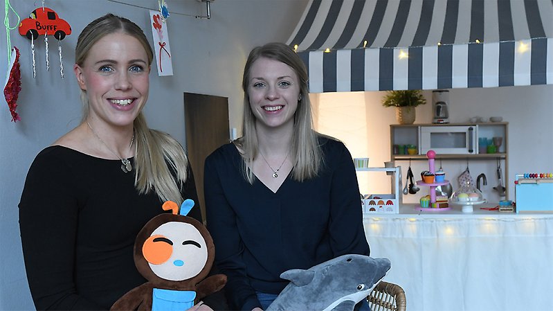Två unga kvinnor tittar in i kameran. Bakom dem finns leksaker.