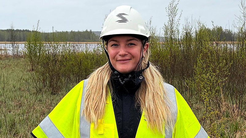 Ung kvinna med varselväst och hjälm står i naturen.