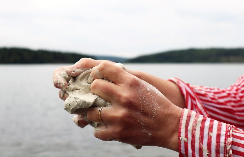 En person ned röd- och vitrandig skjorta håller lera i händerna med en sjö i bakgrunden.