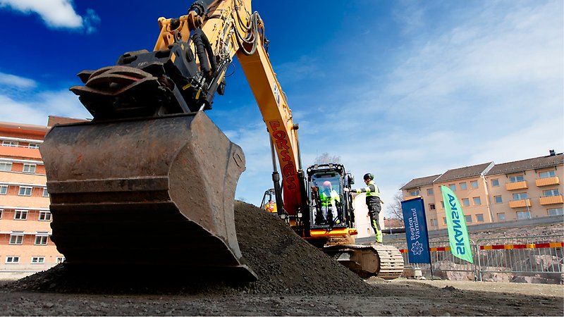 Grävmaskin som tar ett symboliskt första grävtag i grushög. 
