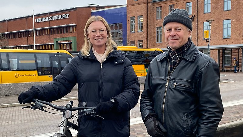 En kvinna och en man står utomhus och tittar in i kameran. Kvinnan håller i en cykel. Bakom dem syns gula bussar.och en byggnad med texten Centralsjukhuset.