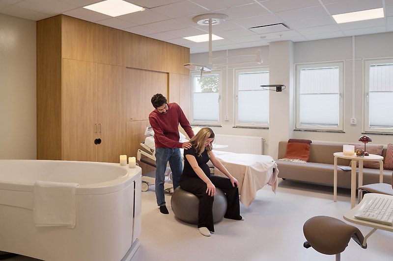 Kvinna sitter på en medicinboll och en man står bakom och håller henne om axlarna.