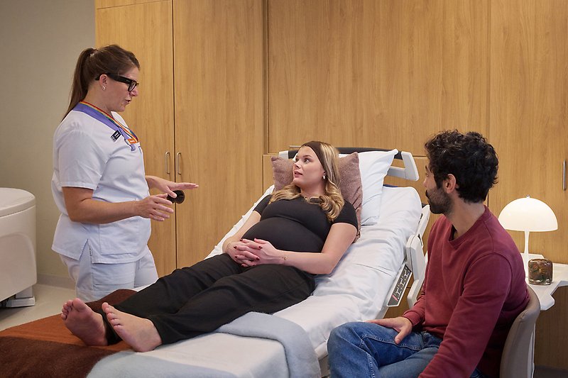 Gravid kvinna i sjukhussäng, man på ena sidan och sjukvårdsperson på andra.